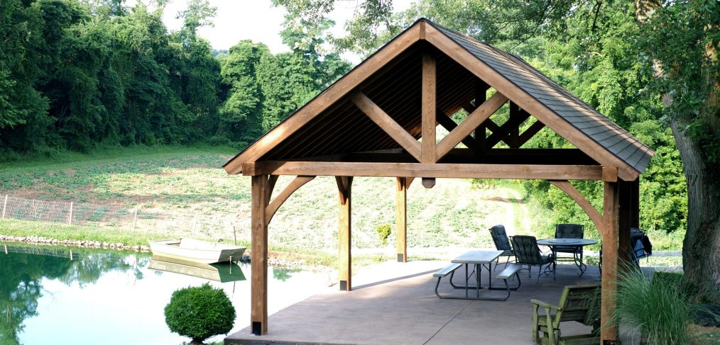 18x32 Open A-Frame Pavilion made with timberwood and asphalt roofing stands next to a pond with tables and chairs underneath it.